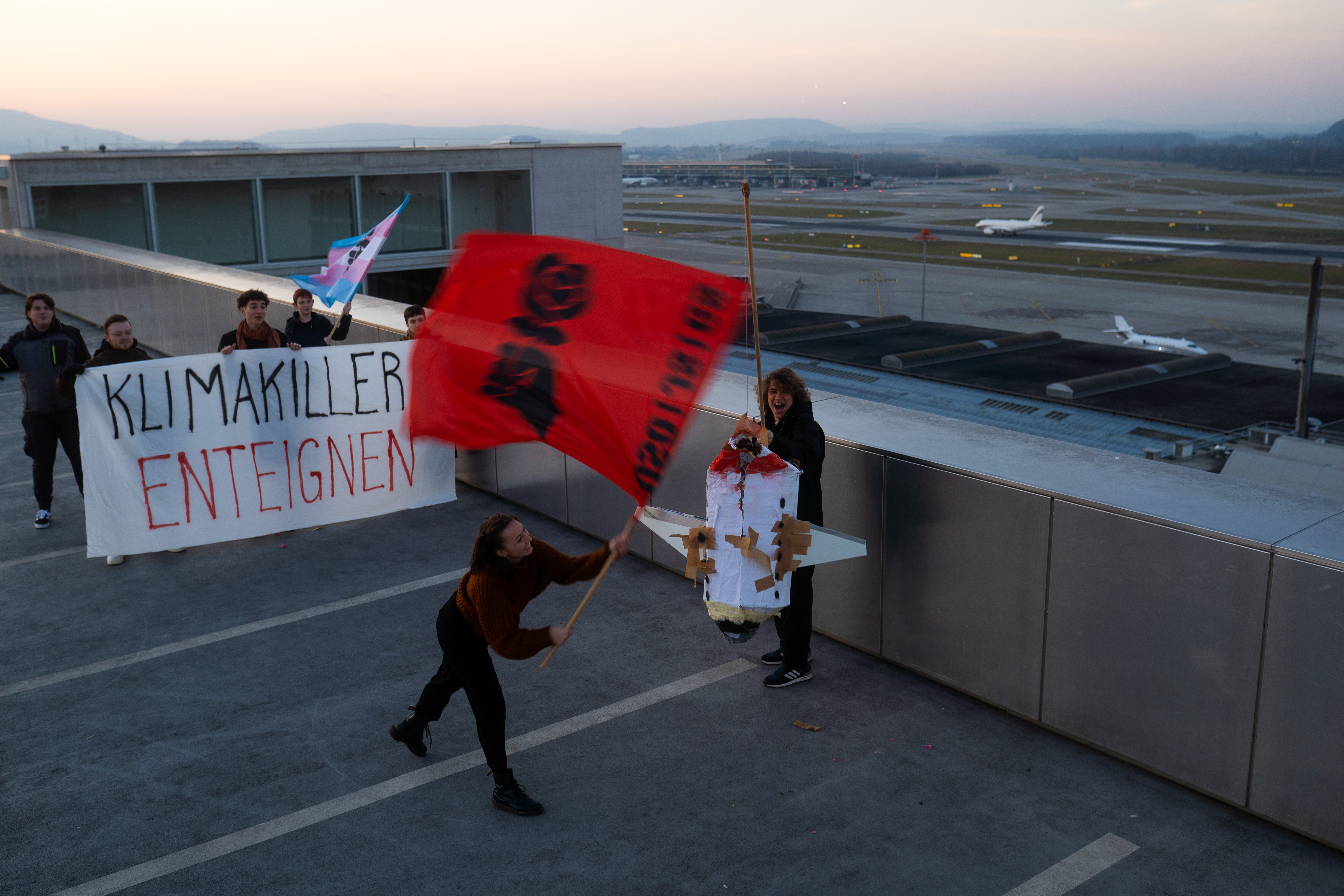 Aktivist*innen der JUSO zerschlagen beim Flughafen Zürich ein Flugzeug, aus dem rote Blüten fallen. Diese stehen für das gute Leben für alle, das nur möglich ist, wenn wir die Kapitalismus überwinden.